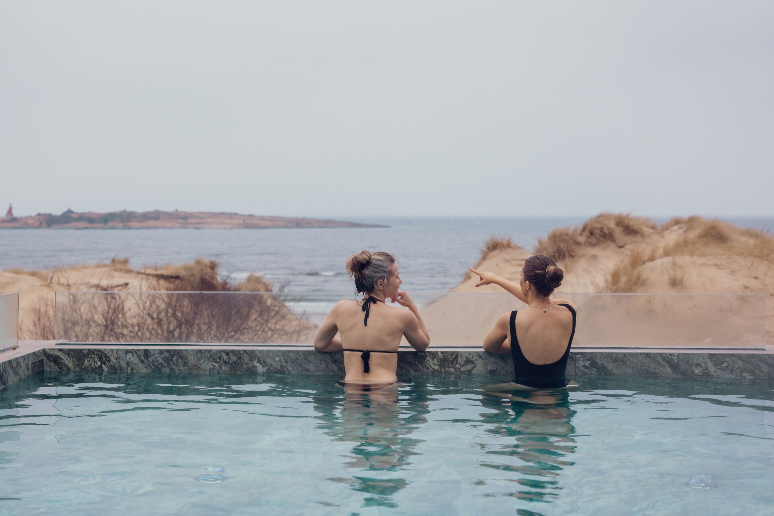 Två kvinnor badar i en pool och blickar ut över havet.